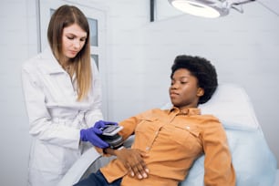 Prevención del cáncer de piel y el melanoma. El médico dermatólogo profesional examina los lunares del paciente en el brazo de la dama africana con la ayuda de un dispositivo moderno para la dematoscopia