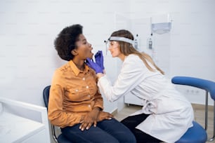 Medico otorinolaringoiatra femminile che esamina la bocca del paziente in una clinica moderna. Medico in ospedale che controlla il mal di gola e le tonsille di una giovane paziente afroamericana