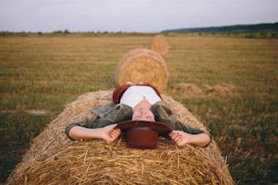 Beautiful carefree woman in hat lying on haystack enjoying evening in summer field. Young happy female relaxing on hay bale in countryside. Atmospheric tranquil moment