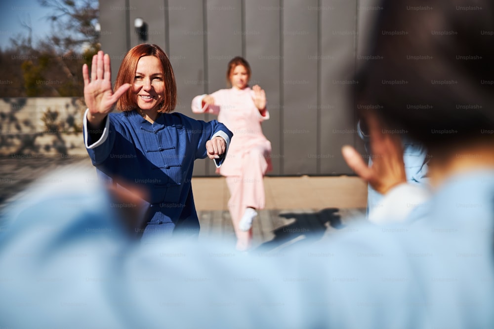 Gladsome lady smiling while practicing tai chi outdoors with her group and master