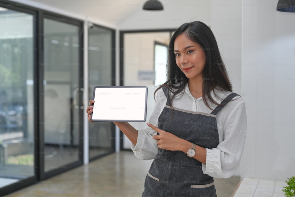 Friendly young woman barista holding and showing digital tablet with empty screen.