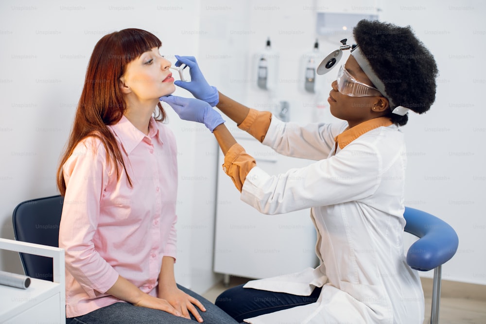 Young woman patient at the modern medical office. Professional African woman doctor otorhinolaryngologist in medical coat and with frontal reflector and eyeglasses checks the woman's nose