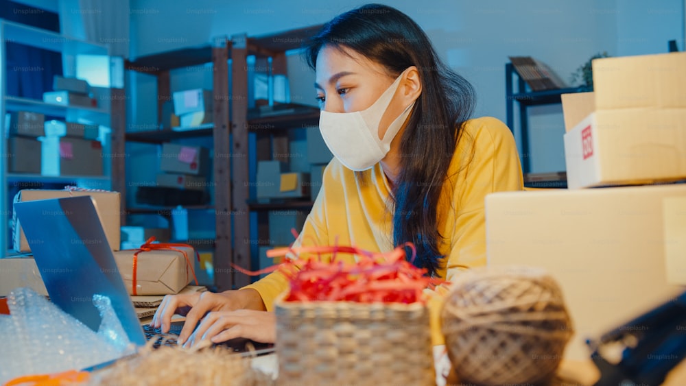 Junge asiatische Geschäftsfrau trägt Gesichtsmaske Bestellung prüfen und Kunden-Posteingang Chat auf Laptop im Home Office in der Nacht. Kleinunternehmer, Online-Marktlieferung, Lifestyle-Freelance-Konzept.