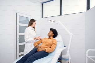 Young African American woman at beauty cosmetology clinic sitting on medical chair, while female doctor wearing white uniform explaining skin treatment procedures