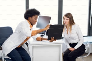 African American female doctor and pretty Caucasian woman patient Discussing the way of treatment or medical record data at modern diagnostic center. Doctor shows a clipboard to patient