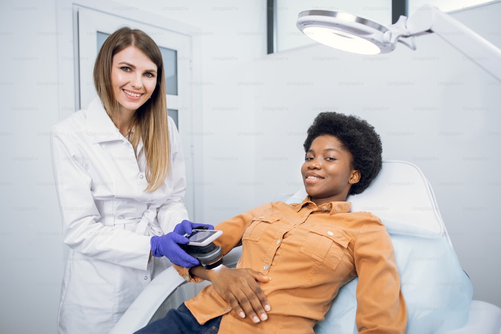 Oncology, skin cancer detection. Pleasant young woman, doctor dermatologist looking at camera with smile, while checking female black patient's mole on hand with modern digital magnifier dermatoscope
