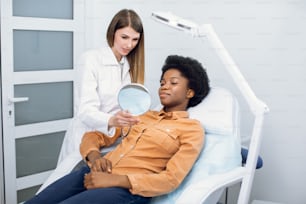 Young beautiful African woman sitting in armchair and looking to mirror in cosmetology clinic. Smiling likable professional beautician explaining skin procedures before mesotherapy or skin treatment