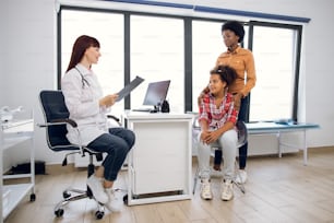 Medicine, healthcare, pediatry and people concept. Afroamerican woman with her cute 12-aged daughter at the appointment at modern medical center. Female friendly doctor talking with patients