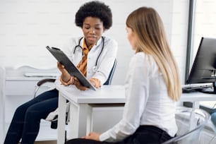 African female physician and young girl patient discussing something while sitting at the table. Medicine and health care concept. Doctor shows clipboard with information for patient
