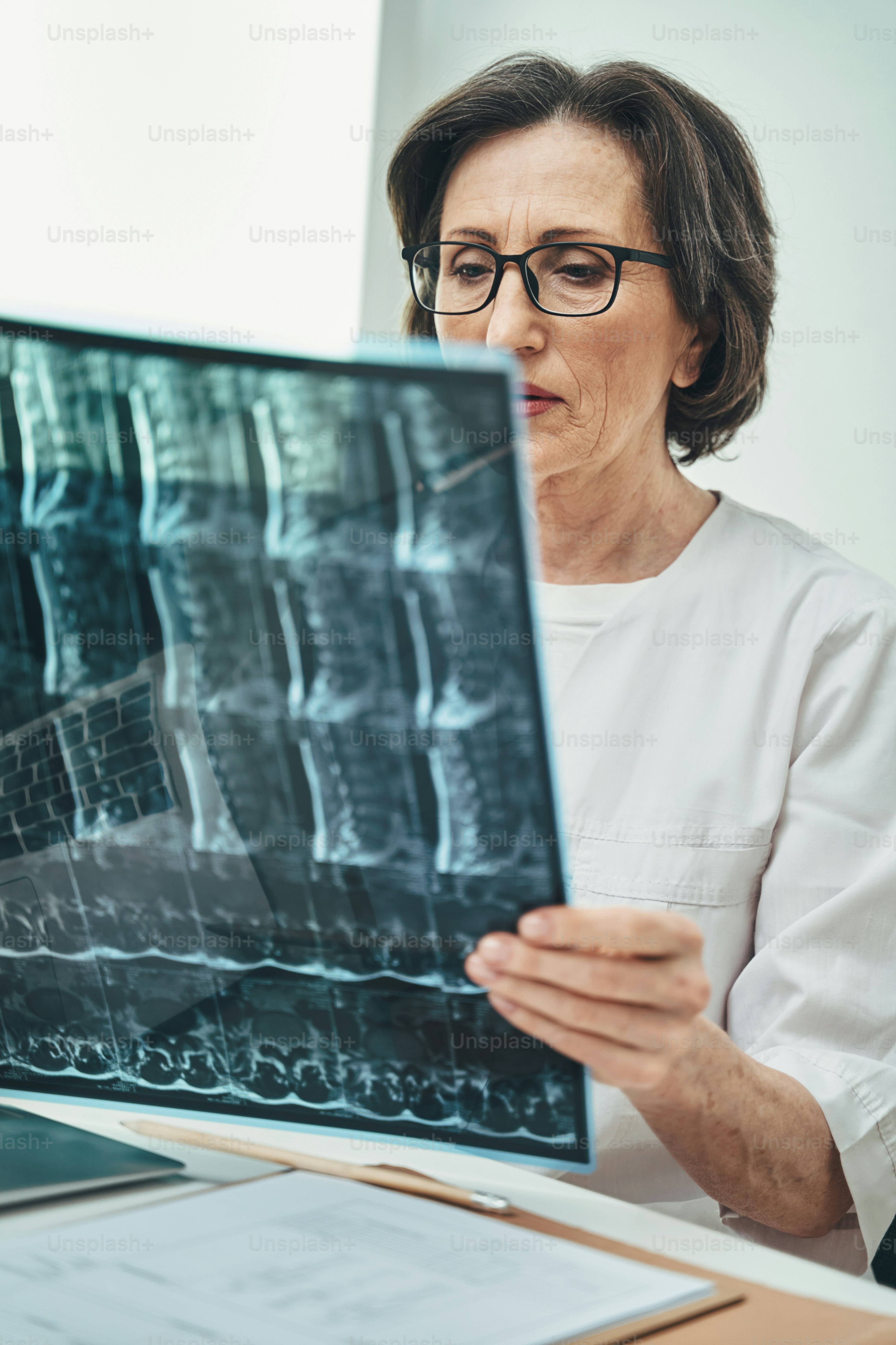 Focused serious mature Caucasian female vertebrologist in eyeglasses sitting at the table in her private office