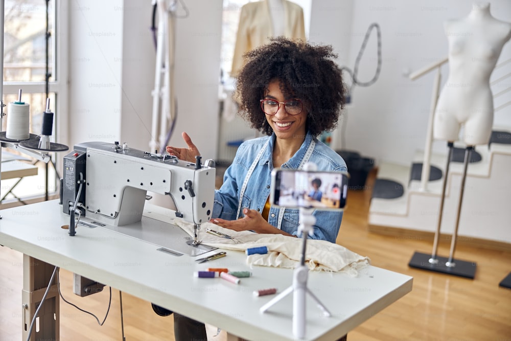 Cheerful African-American lady fashion designer shows contemporary sewing machine shooting new video for blog in light workshop
