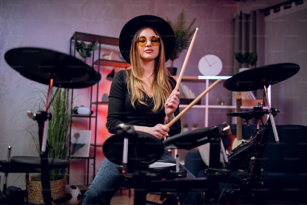 Beautiful young woman in black hat and eyeglasses playing electronic drums indoors. Female musician with long hair rehearsing at studio before concert.