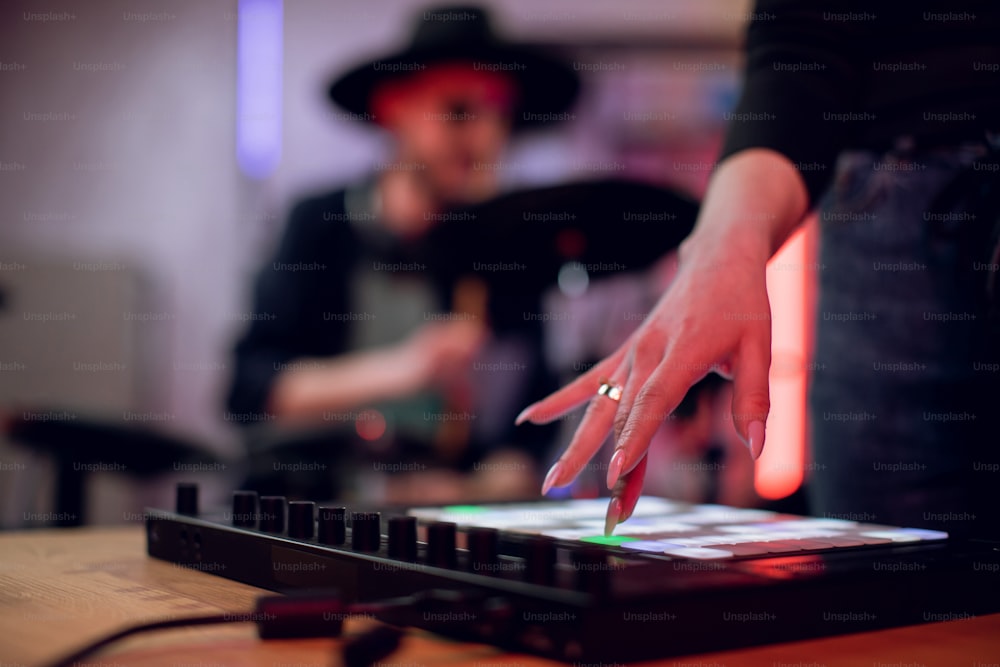 Gros plan d��’une main féminine touchant la table de mixage pour la création d’une chanson à succès en studio. Flou d’arrière-plan d’un artiste masculin jouant sur une batterie électronique. Enregistrement de musique.