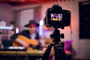 Modern digital camera with handsome musician on screen that playing guitar. Young stylish man sitting at studio and recording online lessons about music.