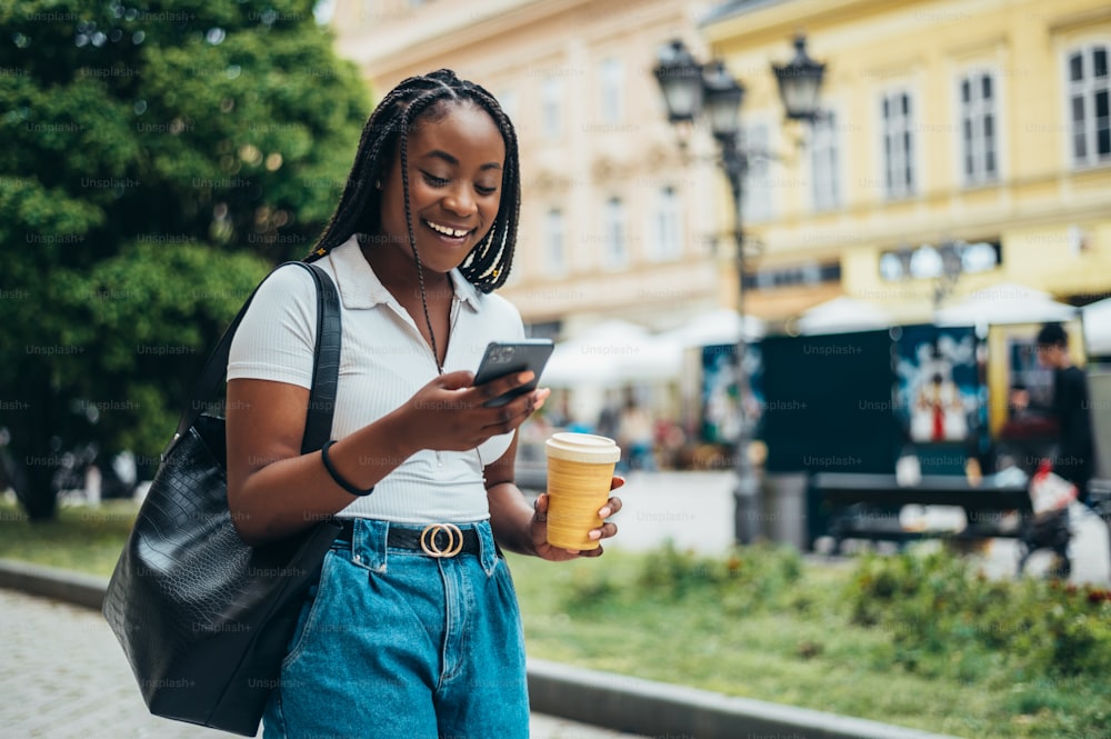 Cheerful african american woman using smartphone and drinking coffee takeaway while out in the city
