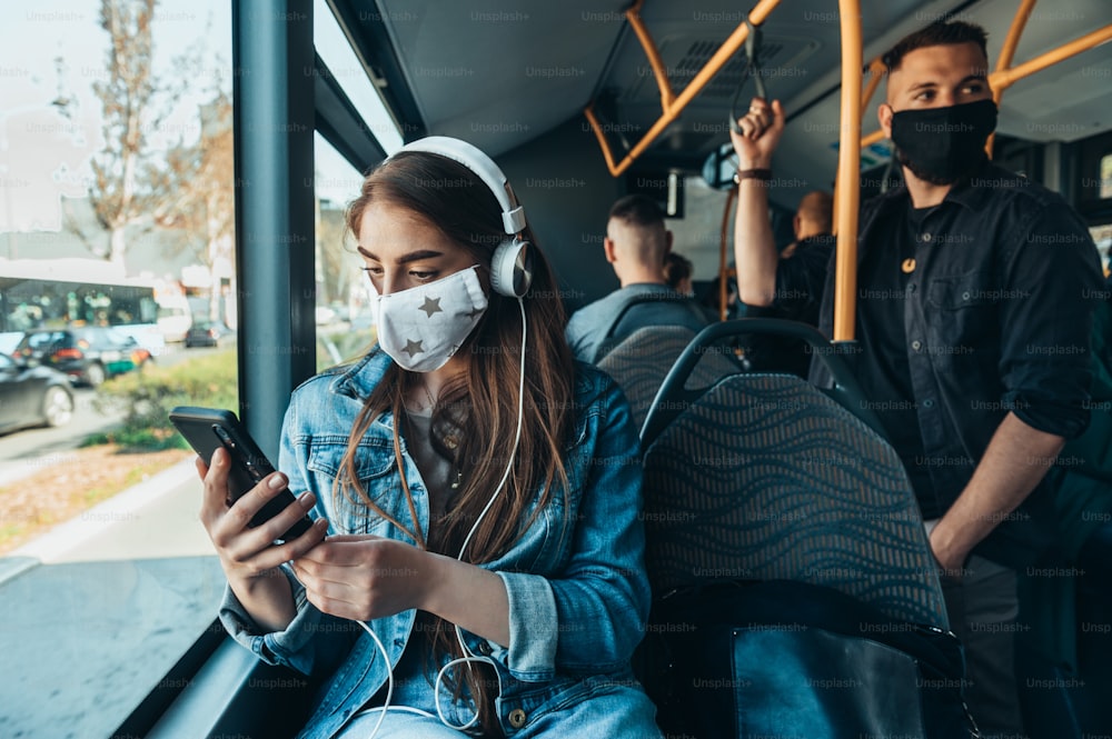 Mujer con máscara protectora mientras viaja en un autobús mientras usa un teléfono inteligente y está sentada a una distancia de otros pasajeros debido a la pandemia de covid19