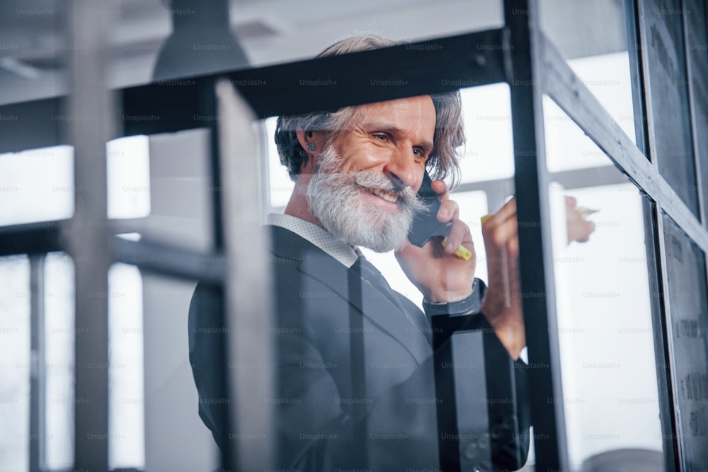 Mature businessman with grey hair and beard in formal clothes have conversation by the phone in the office.