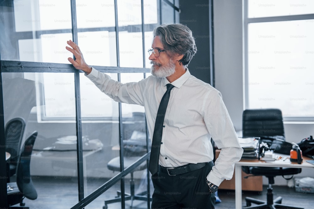 Mature businessman with grey hair and beard in formal clothes is in the office touching glass.
