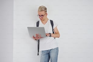 Student in casual clothes and with backpack stands indoors against white wall with laptop.