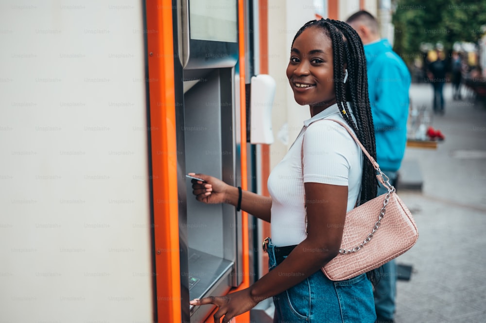 Fröhliche afroamerikanische Frau, die Kreditkarte benutzt und Bargeld am Geldautomaten abhebt