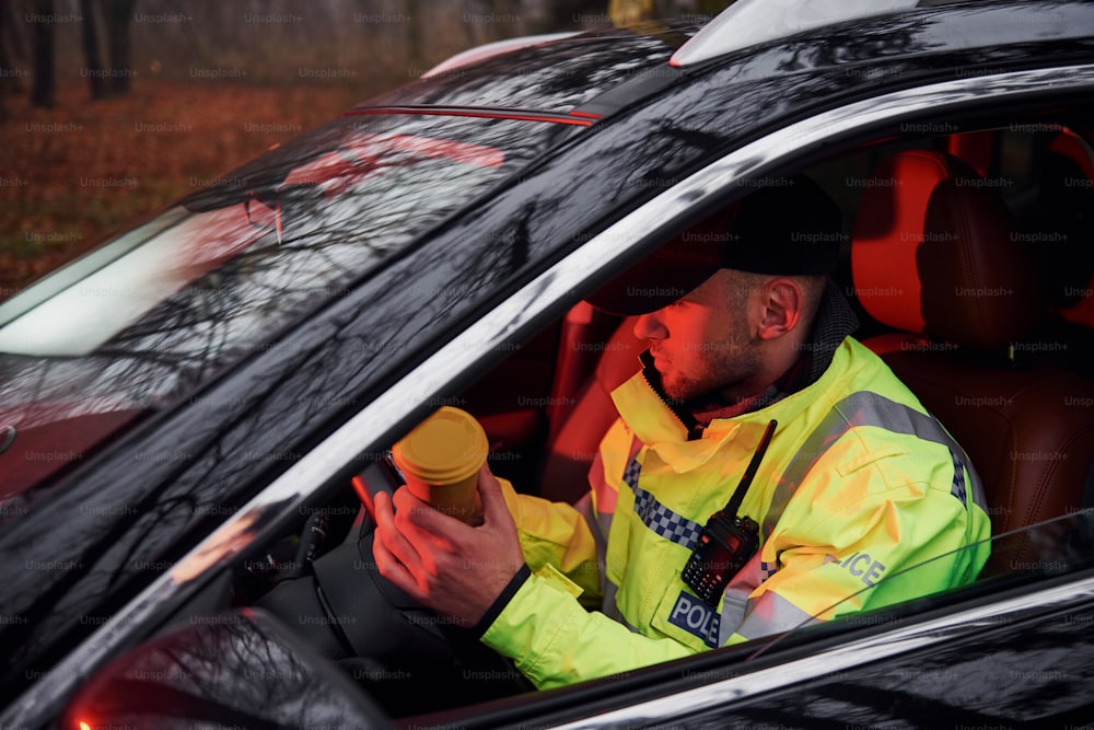 Policial do sexo masculino em uniforme verde sentado em automóvel com copo de bebida.
