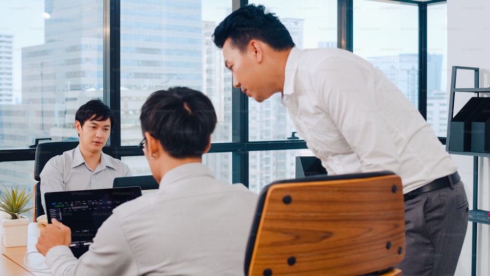 Millennial-Gruppe junger asiatischer Geschäftsleute in einem kleinen, modernen Büro. Japanischer männlicher Chef Vorgesetzter unterrichtet Praktikant oder neuer Mitarbeiter Chinesischer junger Kerl, der bei schwierigen Aufgaben im Besprechungsraum hilft.