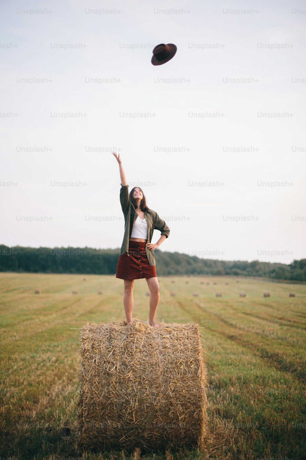 Beautiful carefree woman throw up hat in the sky while standing on haystack in evening summer field. Happiness. Young happy female  having fun on hay bales in countryside. Atmospheric moment