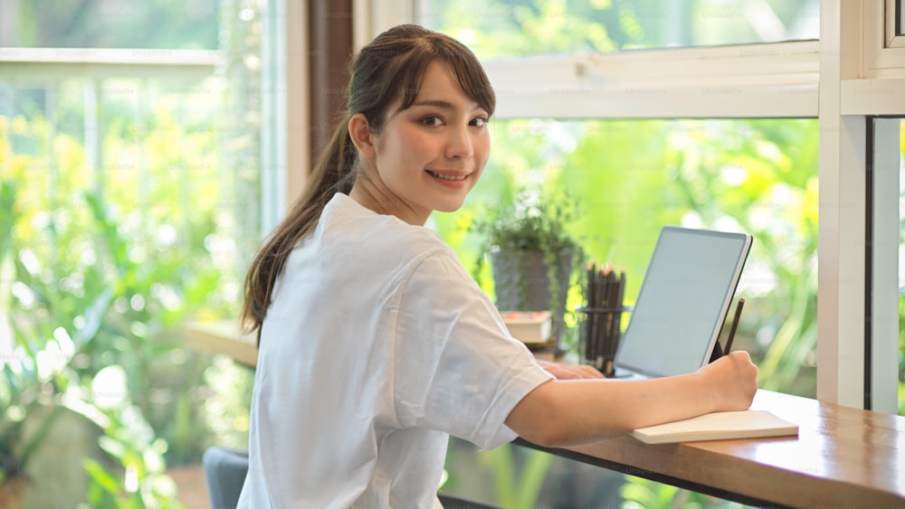 Portrait of young female teenager looking and smiling to camera while working with stationery and tablet