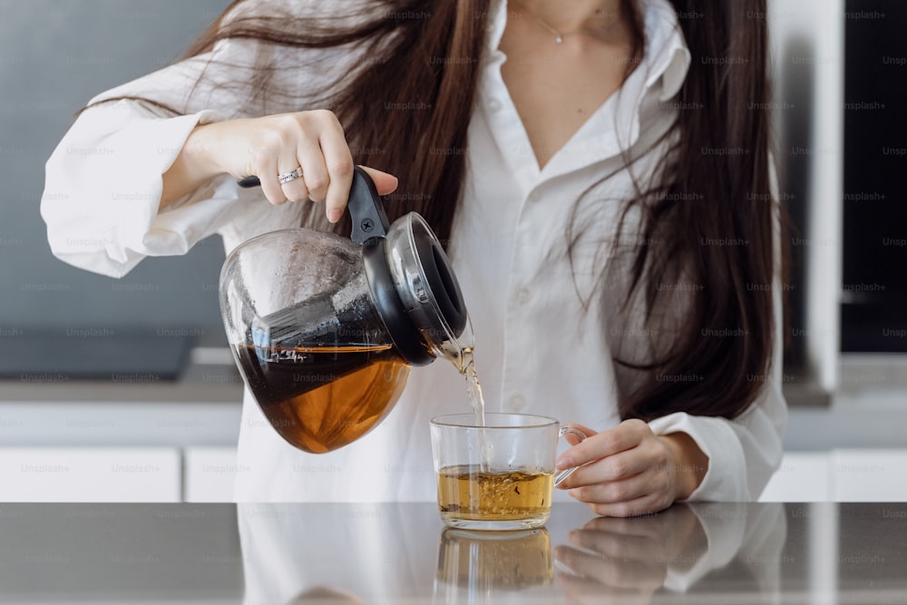 Cropped view of young woman spending morning standing on kitchen, pouring traditional tea from transparent teapot into a glass cup. Female make hot weight loss drink with organic herbs
