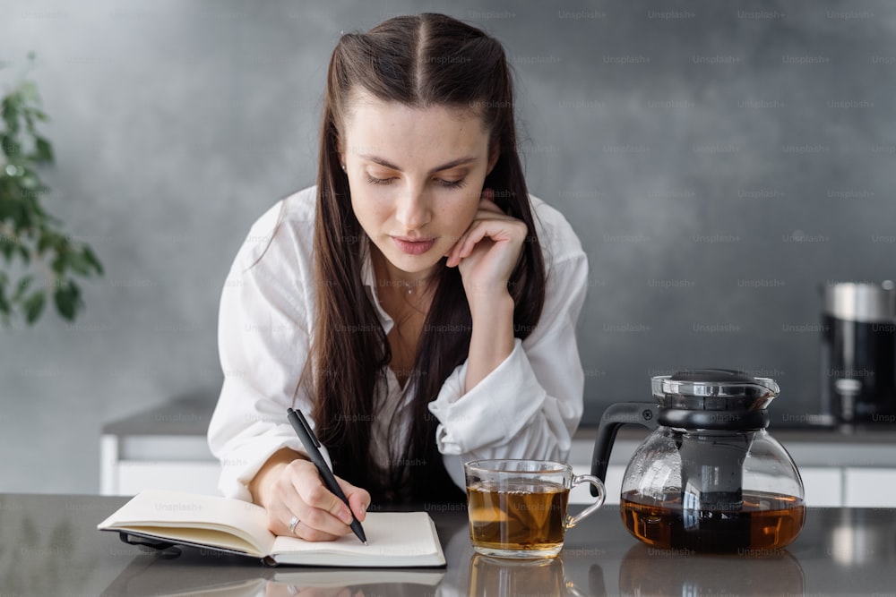 Inspiration and recreation concept. Calm young woman standing behind kitchen table with tea cup, writing in her diary, spend morning at home. Concentrated millennial work with paper notebook plan day