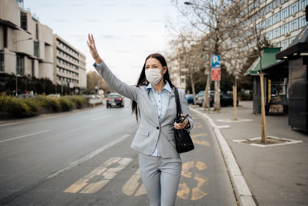 Young and elegant business woman standing on empty city street and wearing protective mask to protect herself from dangerous flu or virus. She waits for transportation on bus shelter. Corona virus or Covid-19 concept.