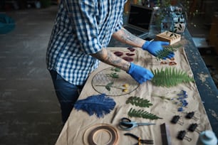 Close up de homem jovem com tatuagens nos braços segurando plantas enquanto está de pé ao lado da mesa com herbário
