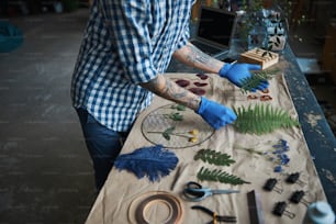 Close up of young man with tattoos on arms holding plants while standing by the table with herbarium