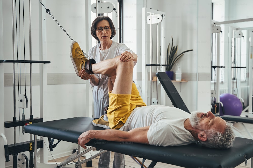 Mature man doing the knee flexion assisted by a qualified physical therapist at the gym