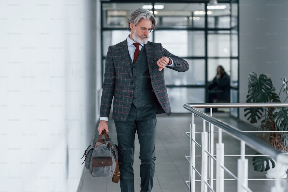 Senior businessman in suit and tie with gray hair and beard walking indoors with bag.