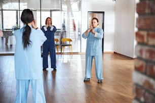 Calm women in loose Chinese suits putting hands to their necks while practicing qigong in a group