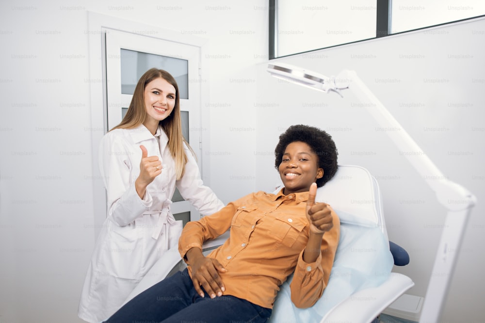 Happy young African American smiling woman, satisfied with results of cosmetic procedures in clinic of esthetic medicine, showing thumb up to camera together with her female doctor cosmetologist