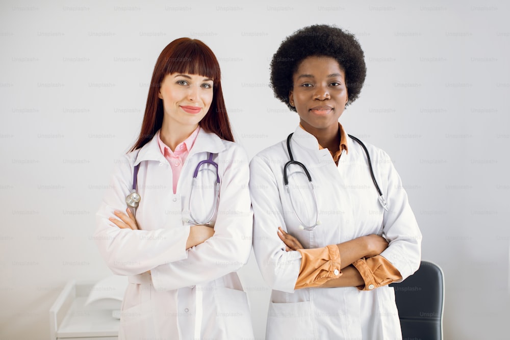 Two smiling happy African and Caucasian female doctors colleagues, wearing white coats, standing side by side with folded arms, posing at camera at modern light medical office