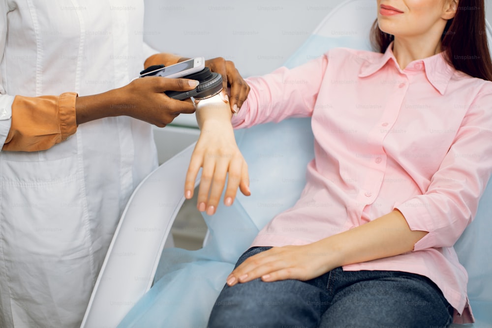 Prevention of melanoma concept. Unrecognizable female afro american doctor examines birthmarks or other benign neoplasms of young lady patient with a special electronic magnifier and phone app