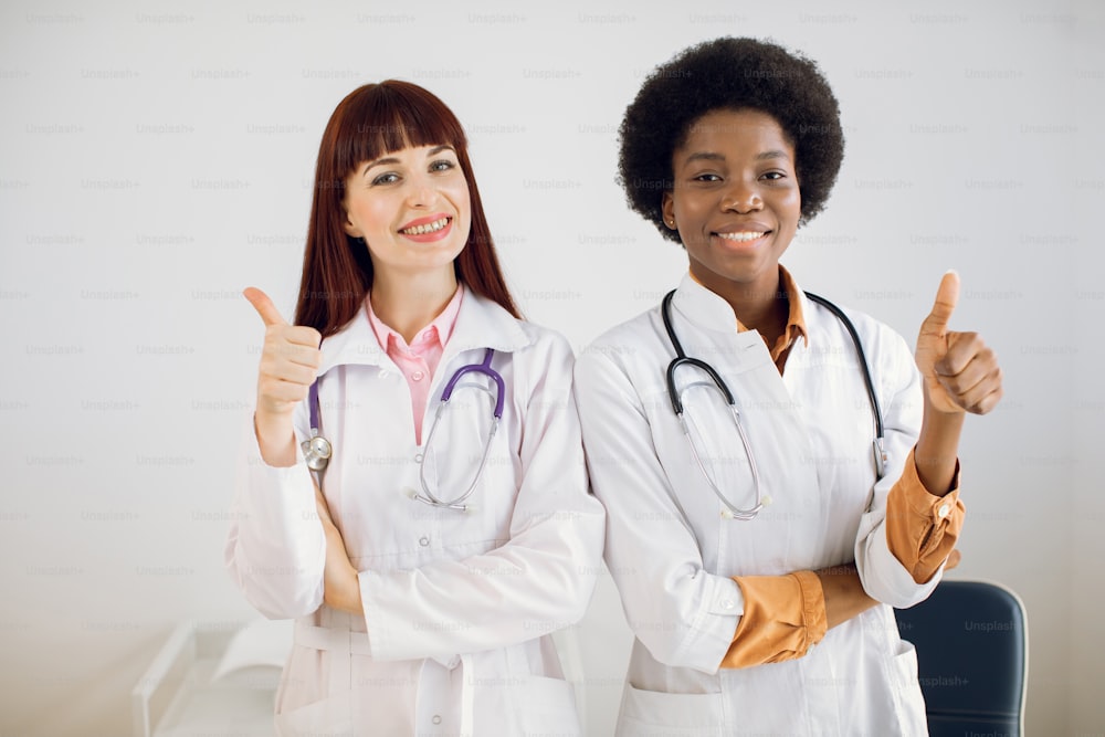 Medical aid, insurance, health care and medicine concept. Two multiracial professional women doctors are standing as a team with thumbs up in a hospital office, smiling at camera