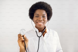 건강 관리 개념, 의료 보험. 의료 사무실에 서 있는 아름다운 미소 짓는 여성 아프리카계 미국인 의사의 초상화, 고립된 흰색 배경에 청진기를 들고 포즈를 취하고 있습니다. 얼굴에 초점 맞추기