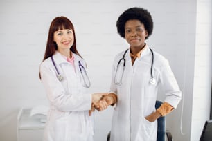 Multiethnic team of doctors. Healthcare and professionalism concept. Cooperation and teamworking. Two diverse confident women doctors, afro american and european, handshaking in the hospital room