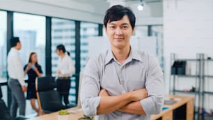 Retrato do empresário executivo bonito de sucesso roupas casuais inteligentes olhando para a câmera e sorrindo, braços cruzados no local de trabalho de escritório moderno. Jovem cara da Ásia em pé na sala de reuniões contemporânea.