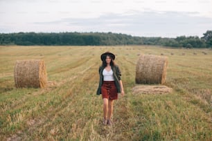Beautiful stylish woman in hat walking at hay bales in summer evening field. Happy young female relaxing at haystacks, vacation in countryside. Tranquility, countryside slow life
