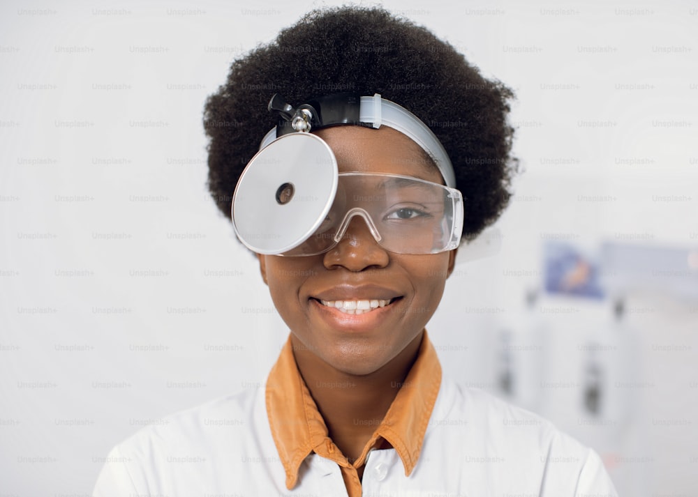 Close up of young friendly smiling African American woman doctor ent otolaryngologist, wearing hed frontal reflector and protective eyeglasses, posing to camera in her modern light office