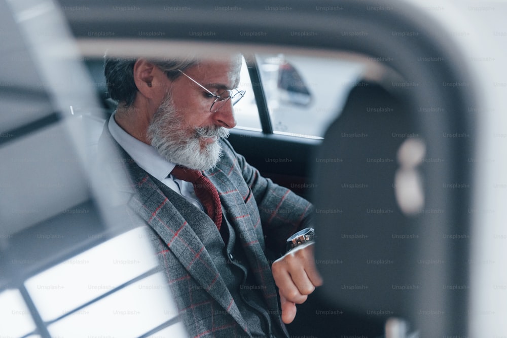 Modern stylish senior man with grey hair and mustache looks at his watches inside automobile.