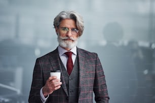 Senior businessman in suit and tie with gray hair and beard standing with cup in hand.