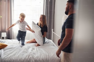 Young married couple with their young son have leisure together in bedroom at daytime.