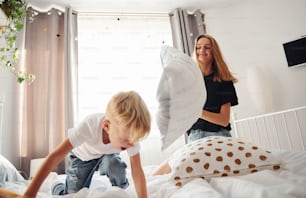 Mother playing pillow fight with her son in bedroom at daytime.