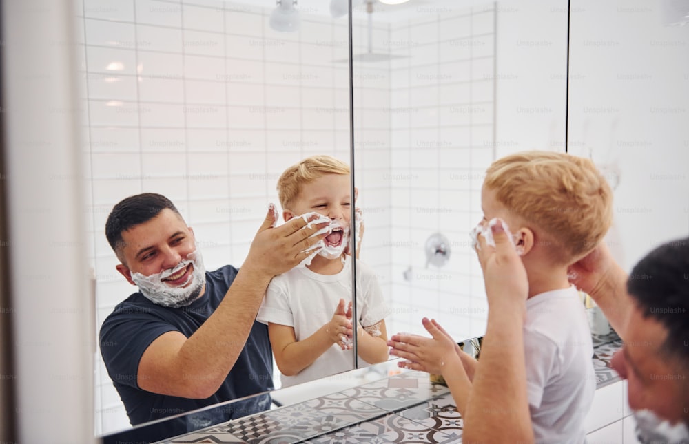 Padre con su hijo está en el baño, diviértete usando gel de afeitar y mirándote en el espejo.